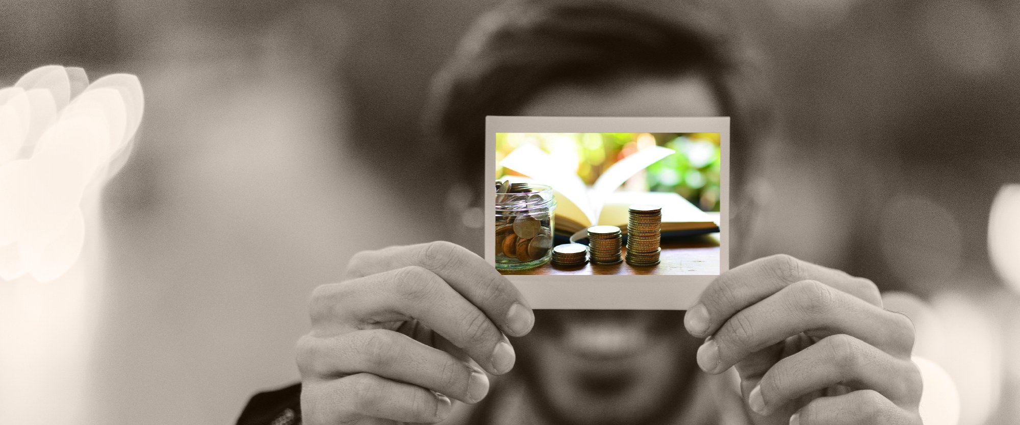 hand holding a polaroid of an image of stacked coins next to a saving jar and in front of an open book